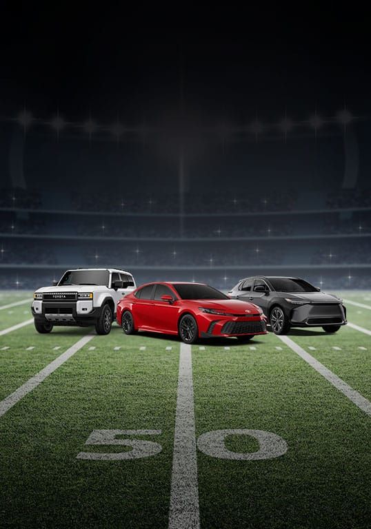 A white Toyota Land Cruiser, a red Toyota Camry, and a gray Toyota bZ4X parked in dynamic angles on a football field under stadium lights.