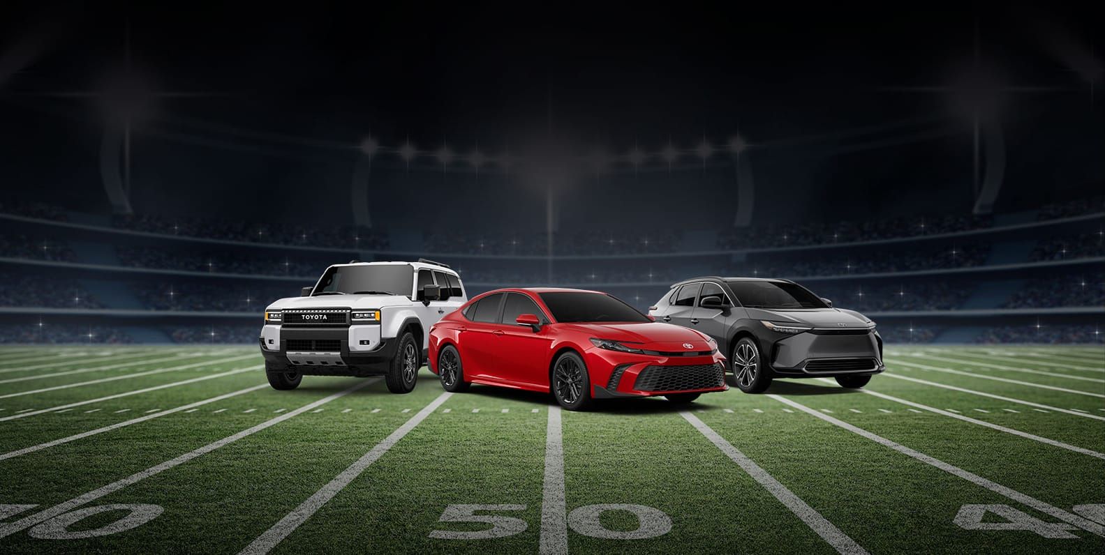 A white Toyota Land Cruiser, a red Toyota Camry, and a gray Toyota bZ4X parked in dynamic angles on a football field under stadium lights.