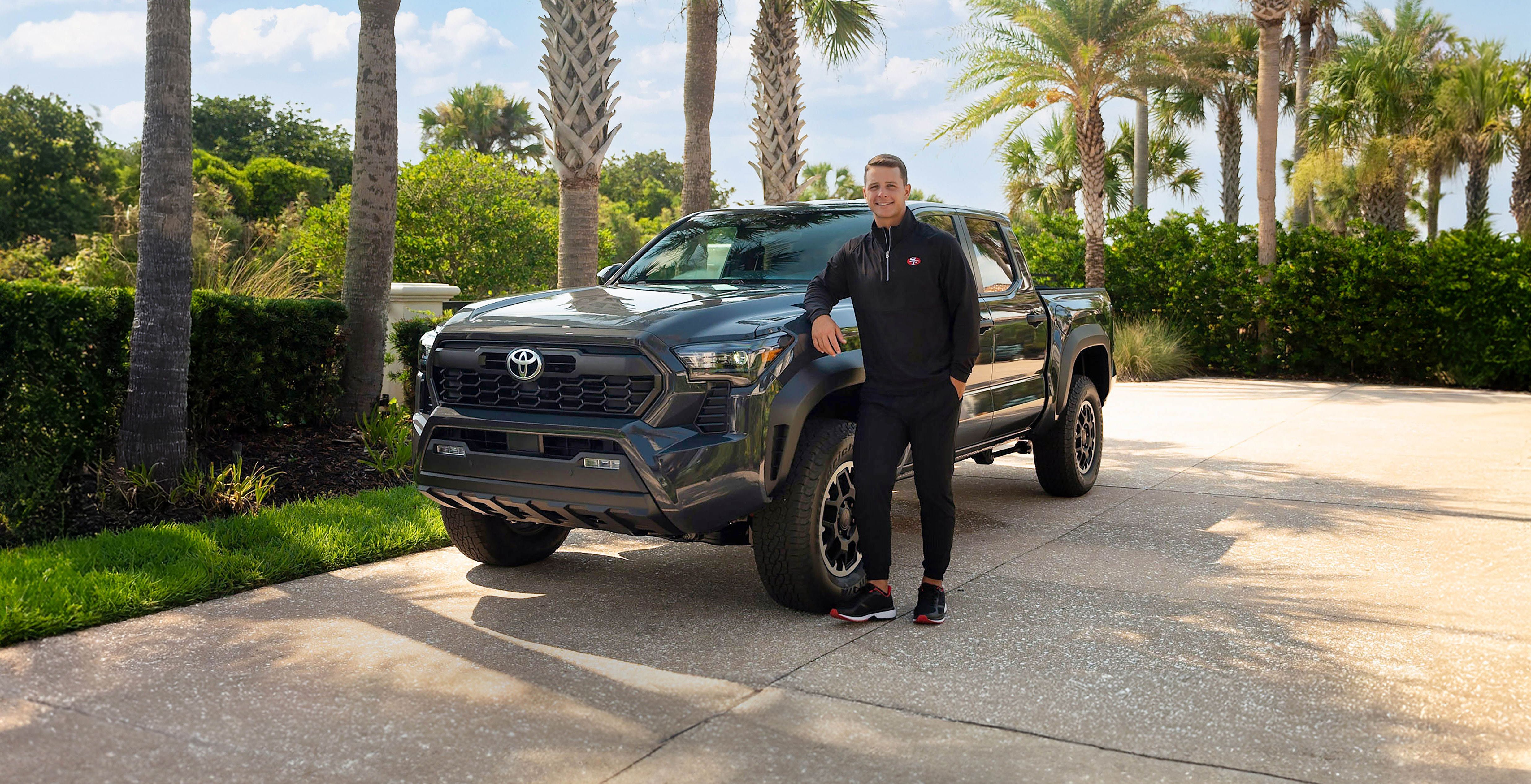 Toyota sponsored athlete, Brock Purdy, in San Francisco 49ers jersey confidently standing next to a gray Toyota Tacoma in a sunny, tropical setting with palm trees, blue sky, and soft white clouds.